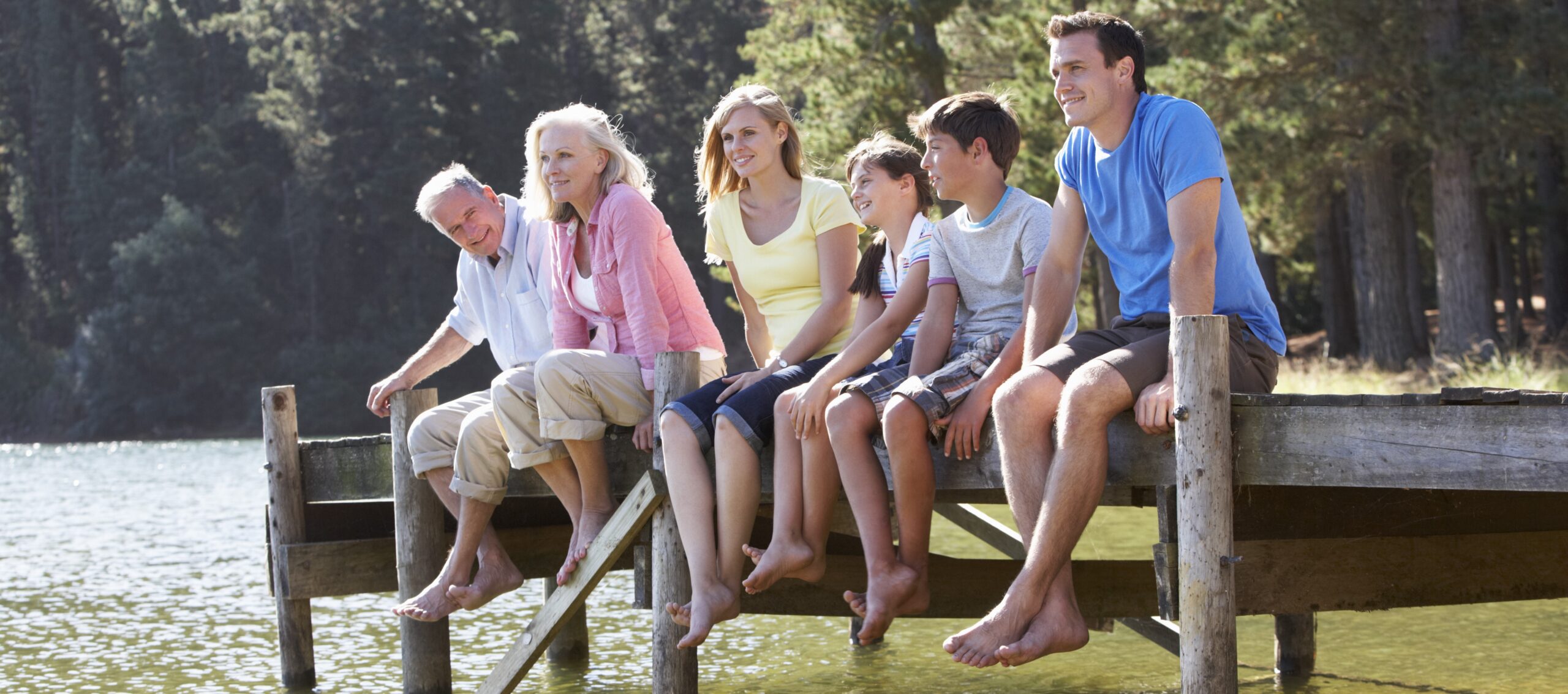Three,Generation,Family,Sitting,On,Wooden,Jetty,Looking,Out,Over